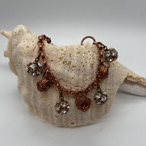 May include: A copper-toned bracelet with multiple charms. The bracelet features small, sparkling crystal ball charms and wire-wrapped accents. The bracelet is displayed on a large, textured seashell against a white background.