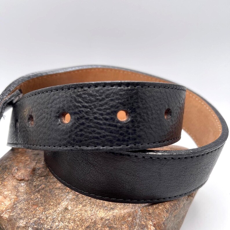 May include: A black leather belt with visible stitching and three evenly spaced holes. The belt's interior is a light brown color, and the leather has a textured appearance. The belt is curved, suggesting it is flexible and designed for wear.