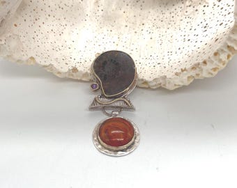 Puede incluir: Un colgante con un fósil de amonita en espiral de color marrón oscuro engastado en plata, acentuado con una pequeña piedra morada y un detalle de alambre dorado. Debajo, una piedra naranja circular está engastada en plata martillada con borde texturizado.