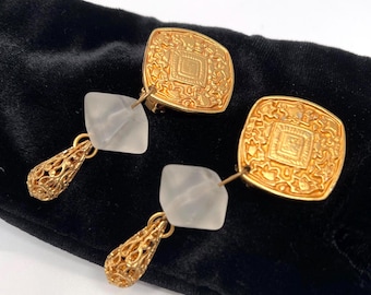 Puede incluir: Un par de pendientes dorados de clip. Cada pendiente presenta una parte superior cuadrada dorada en relieve con un diseño de diamante central. Debajo, una piedra cuadrada esmerilada está conectada a una lágrima de filigrana dorada. Los pendientes se muestran sobre una superficie negra.