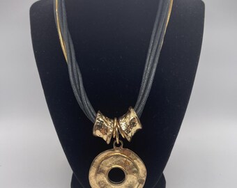Puede incluir: Un collar de múltiples hilos con cordones gris oscuro y una cadena dorada. El colgante consta de dos piezas metálicas doradas texturizadas conectadas a un gran disco circular martillado de color dorado con un orificio central.