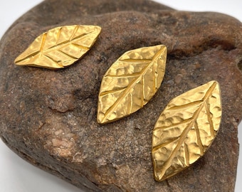Puede incluir: Tres dijes de metal color dorado con forma de hoja y una superficie texturizada y martillada. Cada pieza presenta una vena central y venas laterales más pequeñas grabadas en el metal, creando una apariencia botánica realista.