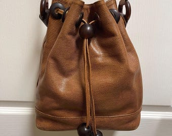 Puede incluir: Un bolso tipo saco de color marrón con un patrón texturizado de piel de lagarto. El bolso cuenta con un cierre de cordón con cuentas de madera marrón oscuro en los extremos. Anillas de madera marrón oscuro unen la correa al cuerpo.