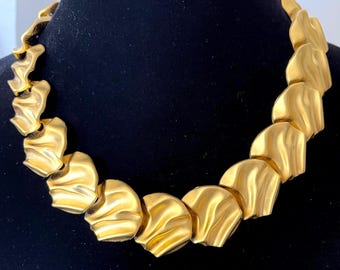 May include: A gold-toned statement necklace with a unique design. The necklace features a series of overlapping, textured, petal-shaped links. The links have a rippled surface, creating a play of light and shadow. The necklace is displayed against a black background.