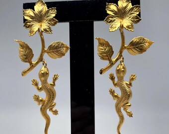 Puede incluir: Un par de pendientes dorados con un diseño de hoja y rama, cada uno con un colgante de lagarto. Los pendientes se exhiben sobre un soporte negro sobre un fondo blanco. El colgante de lagarto cuelga de la rama.