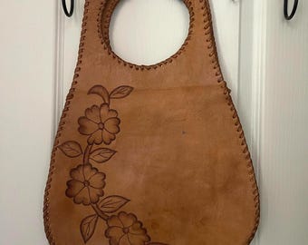 Puede incluir: Un bolso de mano de cuero color canela con un asa circular recortada y un borde con costura de látigo. La parte frontal presenta un diseño floral en relieve de color marrón oscuro con tres flores y varias hojas en un patrón de enredadera.