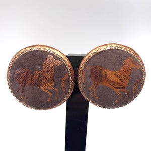 May include: A pair of circular earrings featuring a dark brown textured surface with a metallic copper-colored horse silhouette in the center. The edges of each round earring are framed by a thin border of small, reflective, light-catching crystals.