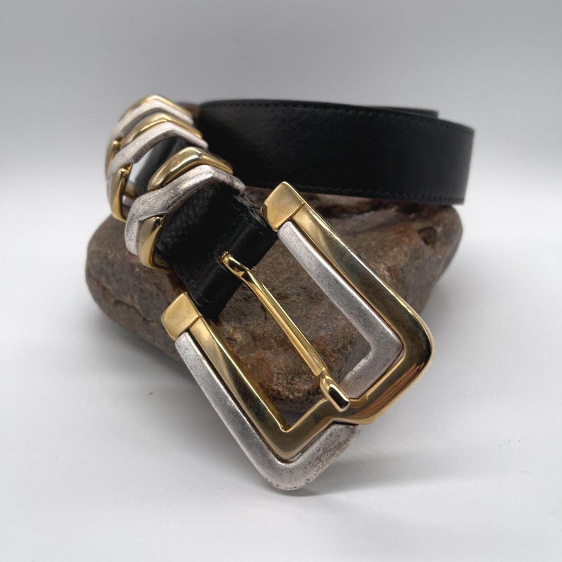 May include: Black leather belt with a silver and gold-tone buckle. The buckle has a rectangular shape with a gold-tone center and silver-tone edges. The belt is resting on a rock.