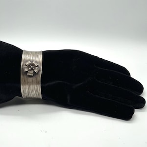 May include: A wide silver cuff bracelet featuring a textured, vertically brushed finish and a central five-petaled flower ornament with a small cluster of silver beads at its core. The metal surface displays a matte, metallic grey appearance.