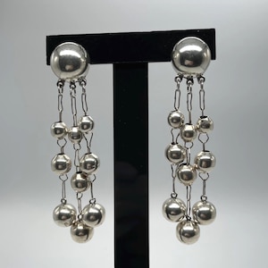 以下が含まれることがあります： シルバーカラーのぶら下がりイヤリング。各イヤリングは、そこから吊り下げられた、卒業したシルバービーズの複数のストランドを備えた、大きな丸いスタッドを備えています。ビーズは繊細なチェーンで接続され、滝の効果を生み出しています。イヤリングは黒いスタンドに展示されています。