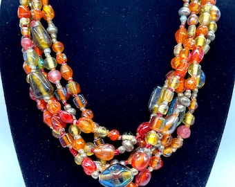 Puede incluir: Un collar de múltiples hilos que presenta un surtido de cuentas de vidrio translúcido en tonos naranja, rojo, amarillo y ámbar, intercaladas con cuentas esféricas metálicas de tono plateado, creando una apariencia texturizada.