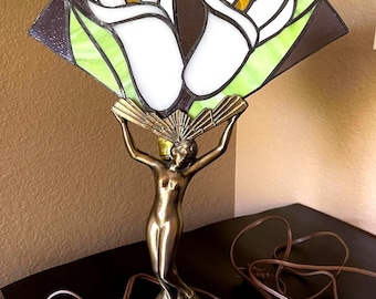 Puede incluir: Una lámpara de mesa con una figura de metal en tono bronce sobre una base circular, sosteniendo una pantalla de vitral en forma de abanico. La pantalla muestra dos flores de cala blancas con centros amarillos sobre un fondo marrón y verde.
