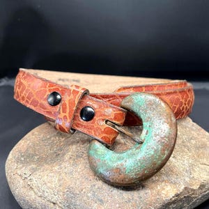 Może przedstawiać: Brązowy skórzany pasek z teksturowanym wzorem i unikalną, postarzaną miedzianą klamrą. Pasek ma czarne guziki i klasyczny design.