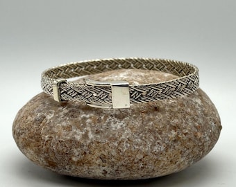 Puede incluir: Una pulsera de tono plateado que presenta un patrón de metal trenzado en toda la banda. La pulsera incluye un mecanismo de cierre metálico rectangular y liso que asegura los extremos, creando un diseño circular continuo.
