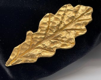 Puede incluir: Un broche de metal dorado con forma de hoja de roble. La superficie de la hoja presenta un acabado texturizado y martillado con venas en relieve que recorren el centro y se extienden hacia los bordes festoneados e irregulares de la hoja.