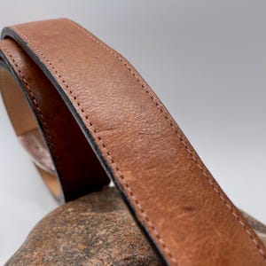 May include: Close-up of a brown leather belt with a stitched edge. The belt is curved, showing the texture and color variations of the leather. The stitching is a lighter color than the belt itself.