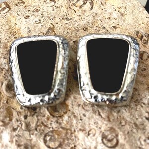 May include: A pair of earrings featuring a central black trapezoidal inlay surrounded by a textured silver-toned border. The earrings have a rounded rectangular shape with a hammered metal finish on the outer edges, creating a distinct contrast between the smooth dark center and the reflective metallic frame.