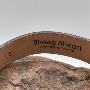 May include: Close-up of a gray leather belt with the text "Streets Ahead, Santa Monica, California" printed on the inside. The belt is size "S". The belt is resting on a textured, brown surface.