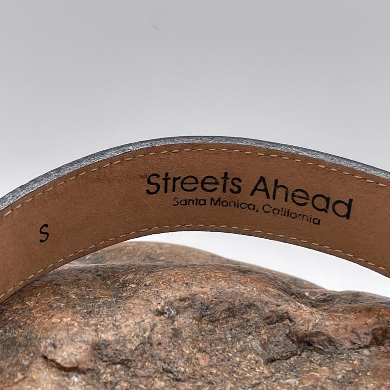 May include: Close-up of a gray leather belt with the text "Streets Ahead, Santa Monica, California" printed on the inside. The belt is size "S". The belt is resting on a textured, brown surface.