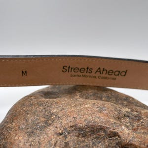 May include: A close-up of a black leather belt with the text "Streets Ahead Santa Monica, California". The belt is size M. The belt is resting on a textured, brown surface.