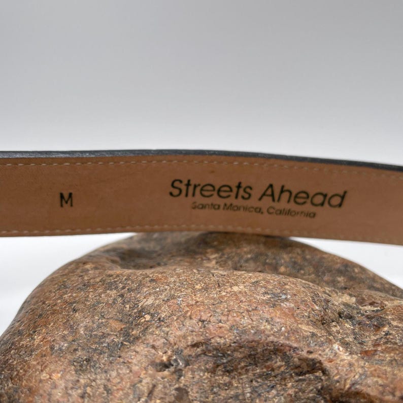 May include: A close-up of a black leather belt with the text "Streets Ahead Santa Monica, California". The belt is size M. The belt is resting on a textured, brown surface.