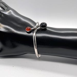 May include: A silver bypass cuff bracelet featuring two spherical terminals. One end is set with a smooth, polished black stone, while the other end is set with a smaller, translucent reddish-orange amber-colored stone.
