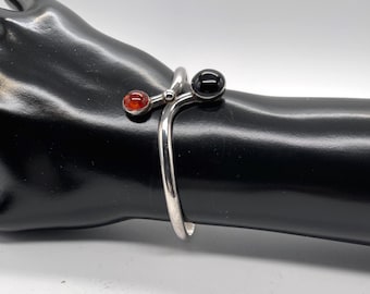 Puede incluir: Un brazalete de plata tipo bypass con dos terminales esféricos. Un extremo está engastado con una piedra negra lisa y pulida, mientras que el otro extremo presenta una piedra más pequeña, translúcida y de color ámbar anaranjado.