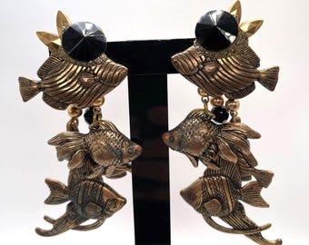 Puede incluir: Un par de pendientes vintage con diseño de pez. Los pendientes son de metal color bronce y presentan tres peces apilados. Cada pendiente tiene un gran cristal negro y detalles dorados.