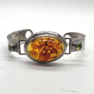 Peut inclure: Un bracelet manchette en argent avec une grande pièce centrale ovale en ambre aux inclusions visibles. Le bracelet est orné de chaque côté d'une petite pierre précieuse ronde et verte sertie dans une monture en argent, reliée à l'ovale central.