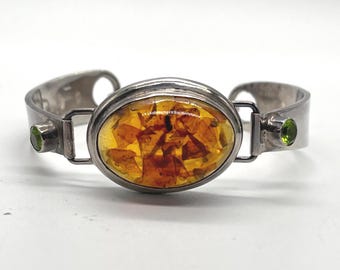 Puede incluir: Un brazalete de plata con una pieza central ovalada de ámbar con inclusiones visibles. El brazalete está acentuado a cada lado por una pequeña piedra preciosa redonda de color verde engastada en plata, conectada al óvalo central.