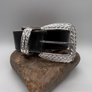 May include: A black leather belt with a silver-colored buckle and accents. The buckle has an ornate, textured design. The belt is displayed on a brown rock against a neutral background.