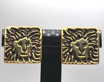 Puede incluir: Un par de pendientes cuadrados dorados con un diseño de cabeza de león. La cara del león está en el centro, rodeada de un fondo oscuro y texturizado. Los pendientes se muestran sobre un soporte negro sobre un fondo blanco.