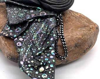 Puede incluir: Broche de cuero negro con un diseño en capas. El broche presenta un elemento circular central con una piedra iridiscente transparente y una cadena decorativa. El cuero tiene un patrón texturizado y moteado con toques de azul, morado y verde.