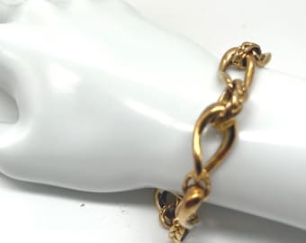 Puede incluir: Una pulsera dorada con un diseño de eslabones de cadena. La pulsera se muestra sobre una superficie blanca. Los eslabones tienen una forma alargada y curvada, y el cierre es visible. La pulsera parece ser de metal.