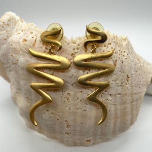 May include: A pair of gold-toned earrings with a unique, wavy design. The earrings have a post-back closure and a matte finish. The earrings are displayed on a beige seashell.