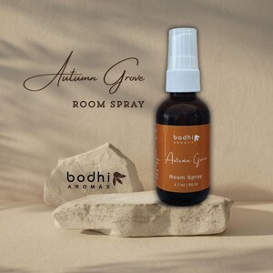 May include: A brown glass bottle with a white spray nozzle, labelled "Autumn Grove Room Spray" and "bodhi AROMAS". The bottle is sitting on a light brown rock.