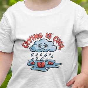 May include: A young child wearing a white t-shirt with the phrase "Crying is Cool" printed on it. The graphic features a crying cloud, raindrops, and broken hearts. The child is smiling and standing outdoors.