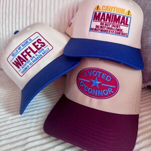 Puede incluir: Tres gorras de b&eacute;isbol con dise&ntilde;os &uacute;nicos. Una gorra tiene el texto "HELLO MY NAME IS WAFFLES" con una visera azul. Otra gorra tiene el texto "CAUTION MANIMAL" con una visera azul. La tercera gorra tiene el texto "I VOTED O'CONNOR" con una visera burdeos.