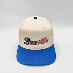 May include: A five-panel baseball cap featuring a cream-colored crown and a vibrant blue flat brim. The front of the cap displays the word Baenitto embroidered in a cursive font with a dark blue outline and a light orange fill color.