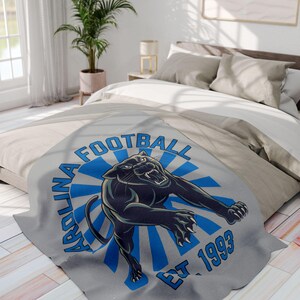 Puede incluir: Una manta de franela gris con un diseño azul y blanco que presenta un pantera negra y el texto "Carolina Football Est 1993".