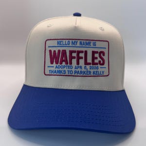 Puede incluir: Gorra de b&eacute;isbol con corona blanca y visera azul. La gorra presenta un dise&ntilde;o de etiqueta con el texto bordado "HELLO MY NAME IS WAFFLES" con la fecha de adopci&oacute;n y un mensaje de agradecimiento.