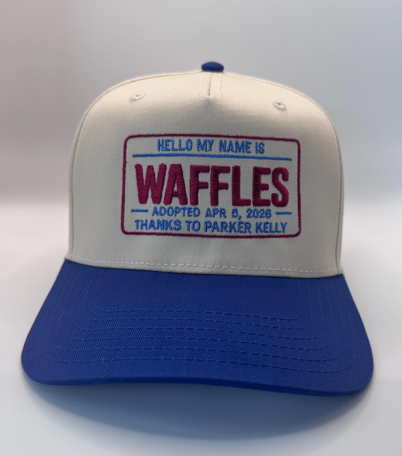 Puede incluir: Gorra de b&eacute;isbol con corona blanca y visera azul. La gorra presenta un dise&ntilde;o de etiqueta con el texto bordado "HELLO MY NAME IS WAFFLES" con la fecha de adopci&oacute;n y un mensaje de agradecimiento.