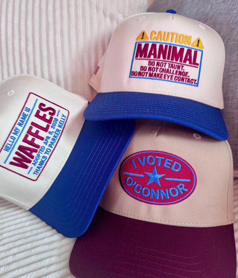 Puede incluir: Una pila de gorras de b&eacute;isbol con varios dise&ntilde;os bordados. Una gorra presenta el texto "CAUTION MANIMAL" con advertencias de seguridad. Otra dice "HELLO MY NAME IS WAFFLES" con detalles de adopci&oacute;n. Una tercera gorra muestra "I VOTED O'CONNOR".