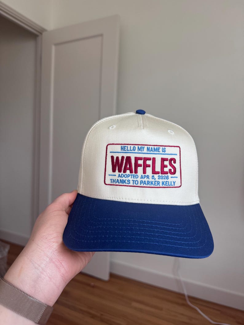 Puede incluir: Gorra de b&eacute;isbol con corona blanca y visera azul. La gorra presenta un parche rectangular con el texto "HELLO MY NAME IS WAFFLES" y detalles de adopci&oacute;n. Un bot&oacute;n azul est&aacute; en la parte superior.
