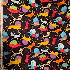Op de afbeelding: Een zwarte stof met een herhalend patroon van kleurrijke cartoonkatten in diverse houdingen, waaronder gele, rode, blauwe en paarse katten, sommige met shirts aan, afgewisseld met effen gekleurde cirkels en tekst langs de witte zelfkant.