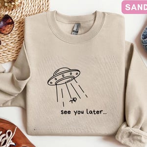 May include: Sand-colored sweatshirt with a crew neck. The front features a black graphic of a UFO beaming up a small figure, with the text "see you later...". The sweatshirt is paired with denim jeans and brown shoes.