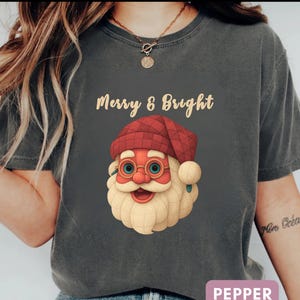 May include: Dark gray t-shirt with the text "Merry & Bright" above a Santa Claus graphic. The Santa graphic features a red hat, glasses, and a white beard. The word "PEPPER" is in a pink rectangle at the bottom.