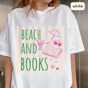 Puede incluir: Camiseta blanca con un diseño de temática playera. El diseño incluye las palabras "BEACH AND BOOKS" en verde, junto con ilustraciones rosas de un cóctel, gafas de sol, un libro abierto y otros elementos de playa.