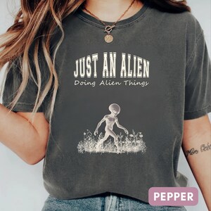 May include: A charcoal gray short-sleeve t-shirt featuring a white graphic of an alien walking through a field of wildflowers. The text reads JUST AN ALIEN in a bold collegiate font, followed by the script text Doing Alien Things below it.