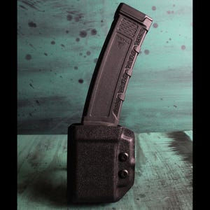 May include: A black magazine and holster set. The magazine is angled and partially inserted into the holster. The holster has a textured surface and two black rivets. The magazine has the text "TRIL" and a triangle logo.
